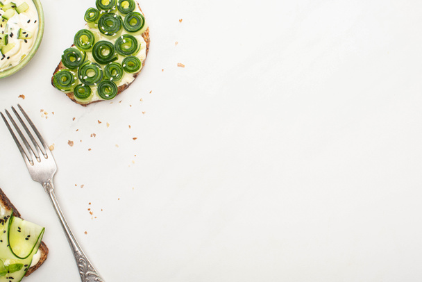 vista dall'alto dei toast di cetrioli freschi con semi vicino allo yogurt in ciotola e forchetta su sfondo bianco
 - Foto, immagini