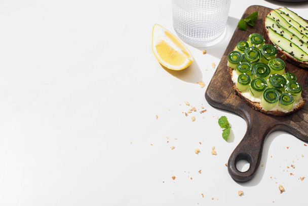 fresh cucumber toasts on wooden cutting board near water and lemon on white background - Фото, зображення