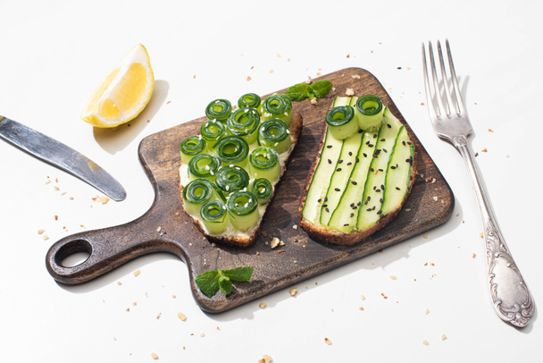 fresh cucumber toasts on wooden cutting board near cutlery and lemon on white background - Foto, Imagen