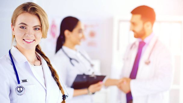 Ελκυστική γυναίκα γιατρός μπροστά από την ιατρική ομάδα, ιατρικό προσωπικό που ελέγχει τα ιατρικά αρχεία στο παρασκήνιο - Φωτογραφία, εικόνα