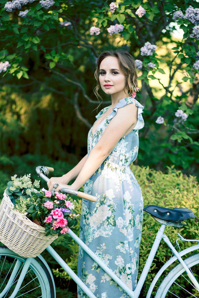 portrait of a beautiful girl in the forest, holding a bike with a basket of flowers, behind the rays of the sun, a blue flowered dress, summer walk relaxation - Фото, изображение