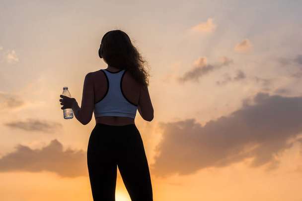 Silhouette young woman in the headphones in the sport clothes with bottle of clear mineral water on the sunset sky backgrounds. The concept of healthy lifestyle - Foto, immagini