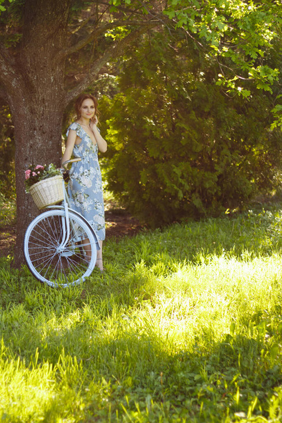 portrait of a beautiful girl in the forest, holding a bike with a basket of flowers, behind the rays of the sun, a blue flowered dress, summer walk relaxation - Photo, Image