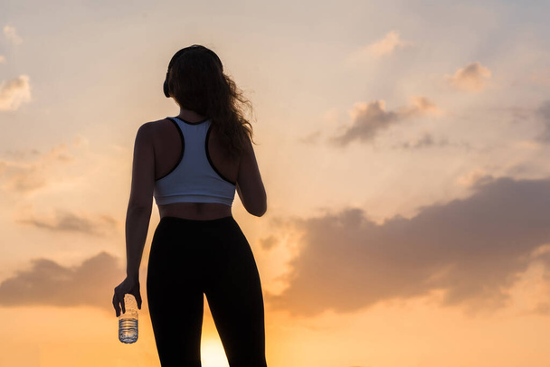 Silhouette young woman in the headphones in the sport clothes with bottle of clear mineral water on the sunset sky backgrounds. The concept of healthy lifestyle - Photo, Image
