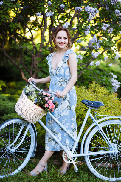 portrait of a beautiful girl in the forest, holding a bike with a basket of flowers, behind the rays of the sun, a blue flowered dress, summer walk relaxation - Valokuva, kuva