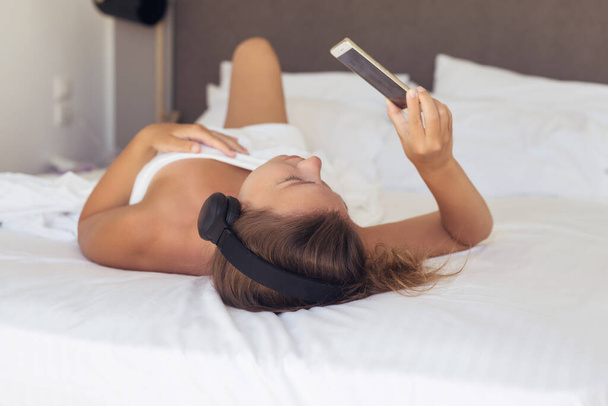 Beautiful woman in headphones listening music and looking at mobile phone lying on a bed in the bedroom and covered sheet. - Foto, afbeelding