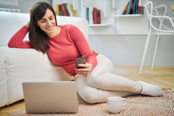 Young smiling woman sitting at home on the floor and using smart phone. - Фото, изображение