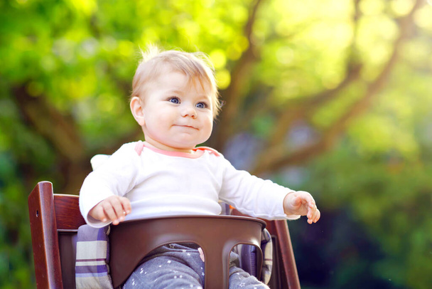 Preciosa adorable niña sentada en la silla alta al aire libre. Beatuiful niño de 6 meses en el jardín en casa, jugando en el día soleado cálido. Bebé sano sonriendo y riendo
. - Foto, imagen