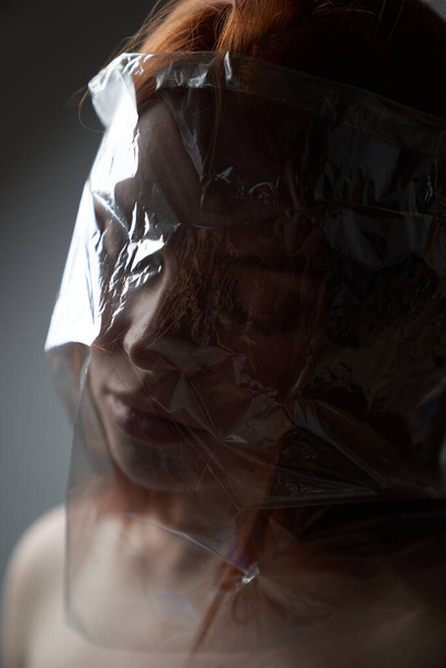 Porträt eines jungen rothaarigen Mädchens auf grauem Hintergrund mit einer Plastiktüte auf dem Kopf. Das Konzept der Plastikverschmutzung der Natur. Überschüssiges Plastik im Leben eines Menschen                                         - Foto, Bild