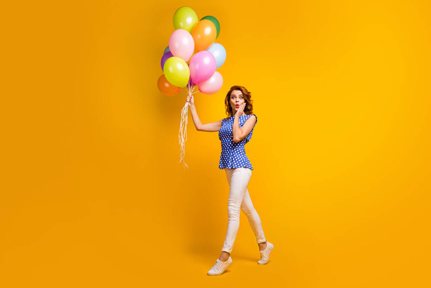 Full body photo of crazy surprised lady hold many colorful balloons moving birthday event hand on cheek wear polka-dot blue shirt white pants shoes isolated yellow color background - Foto, afbeelding