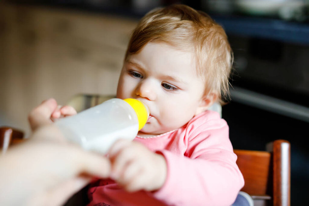Jolie adorable petite fille tenant le biberon et le lait maternisé. Première nourriture pour bébés. Enfant nouveau-né, assis dans la chaise de la cuisine domestique. Bébés en santé et concept d'alimentation au biberon - Photo, image