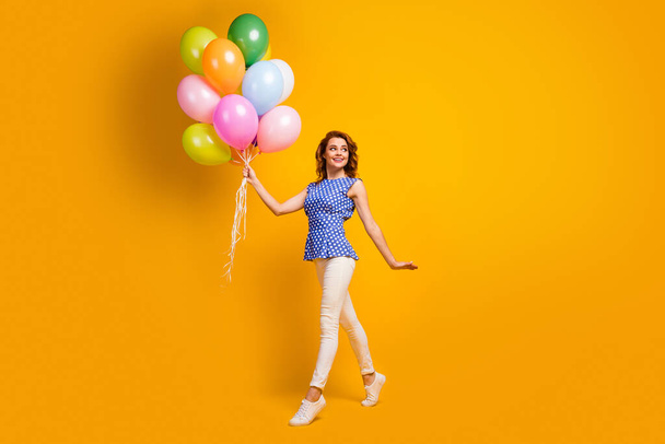 Full body profile photo of funny foxy lady hold many colorful balloons walk street festive mood party wear polka-dot blue shirt white pants shoes isolated yellow color background - 写真・画像