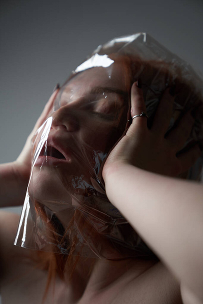   Retrato de una joven pelirroja sobre fondo gris con una bolsa de plástico en la cabeza. El concepto de contaminación plástica de la naturaleza. Exceso de plástico en la vida de una persona
                                       - Foto, Imagen