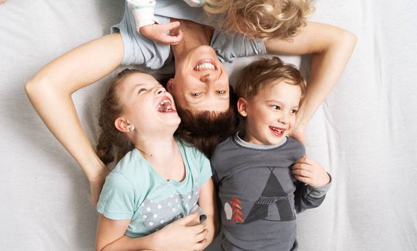 Familie tijd: Mam en drie kinderen liggen vrolijk op een wit bed, knuffelend en lachend. Portret op een witte achtergrond. - Foto, afbeelding