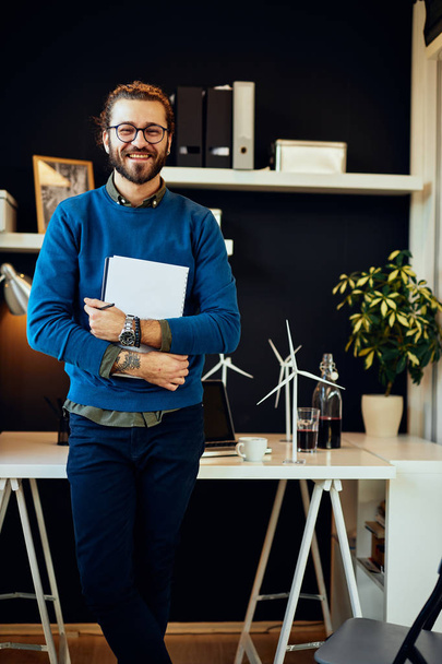 Przystojny młody biały hipster stojący w biurze, trzymający papiery i przygotowujący się do spotkania z ważnymi klientami. - Zdjęcie, obraz
