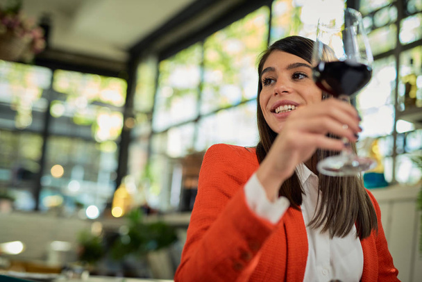 Prachtige glimlachende Kaukasische brunette gekleed elegant zitten in restaurant tijdens lunchpauze en het houden van een glas rode wijn. - Foto, afbeelding