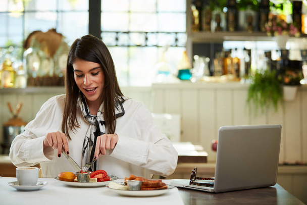 Prachtige Kaukasische elegante brunette zittend in restaurant en worstje etend als ontbijt. Op tafel ligt een laptop. - Foto, afbeelding