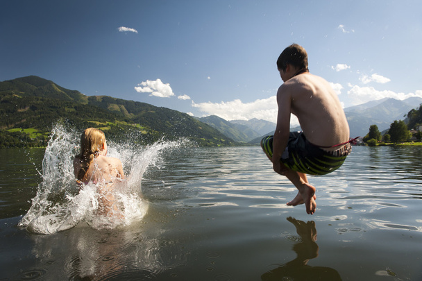 κορίτσι ήδη στο πιτσίλισμα νερού και αγόρι στον αέρα ενώ όπου jumoing σε μια λίμνη, με ωραία φύση και τα βουνά στο βάθος - Φωτογραφία, εικόνα