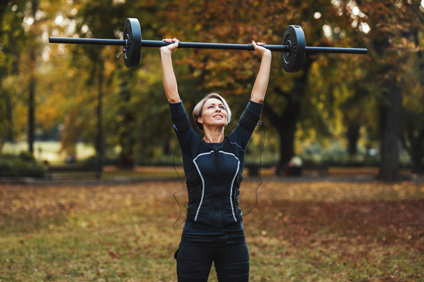 Uma mulher madura muscular está fazendo exercícios com barra no parque, vestida com um terno preto com um simulador eletrônico EMS para estimular seus músculos
. - Foto, Imagem