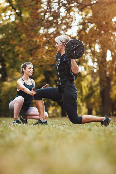 Una donna matura muscolare sta facendo esercizi con personal trainer nel parco, vestita con un abito nero con un simulatore elettronico EMS per stimolare i suoi muscoli
. - Foto, immagini
