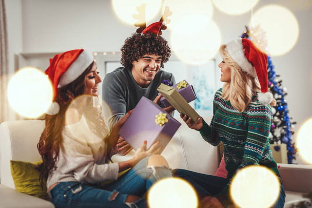 Weseli młodzi przyjaciele ubrani w przytulne ciepłe swetry spędzają czas w salonie na Boże Narodzenie - Sylwester, uśmiechając się, bawiąc i dając sobie prezenty świąteczne. - Zdjęcie, obraz
