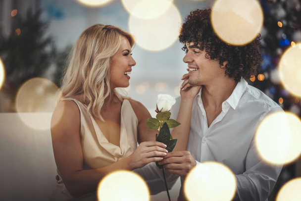 Beautiful young couple is celebrating Christmas at home. Handsome smiling man is giving a white rose to his pretty girlfriend. - Photo, Image