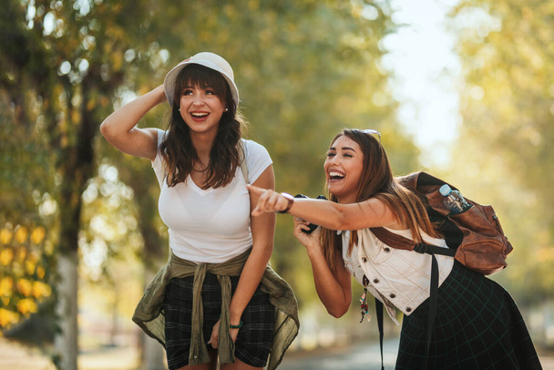 Two beautiful young smiling women with backpacks on their back are walking along the autumn sunny avenue and talking each other and pointing something. - Photo, Image