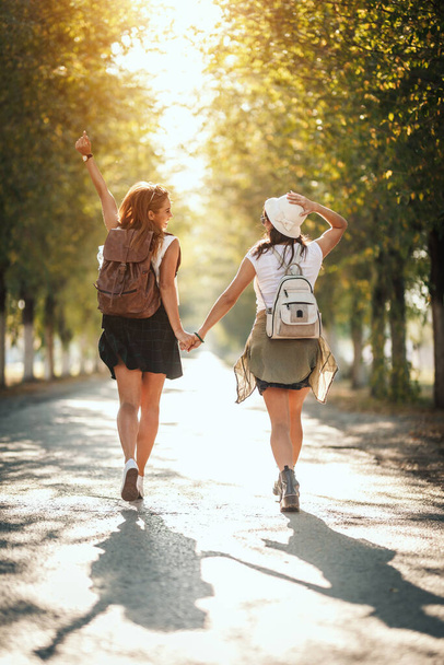 Two beautiful young women with backpacks on their back are walking along the autumn sunny avenue holding hands and looking each other. - Photo, Image