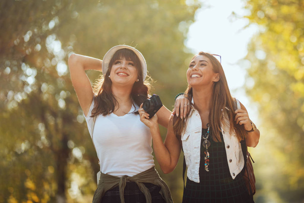 Dvě krásné mladé usměvavé ženy s batohy na zádech kráčejí podzimní slunnou ulicí a dívají se jinam. - Fotografie, Obrázek
