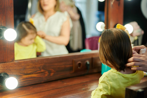 Κοριτσάκι χτένισε τα μαλλιά της μπροστά στον καθρέφτη στο καμαρίνι με τη μαμά. - Φωτογραφία, εικόνα