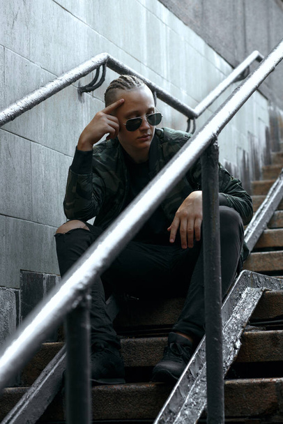 Homem elegante com tranças usa óculos de sol e jaqueta cáqui sentado nas escadas da rua
 - Foto, Imagem