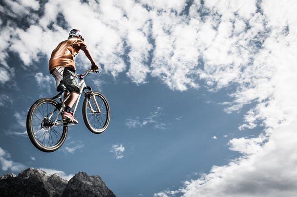 νεαρό αγόρι ψηλά άλματα με το ποδήλατό του μπροστά από τα βουνά και ουρανό - Φωτογραφία, εικόνα