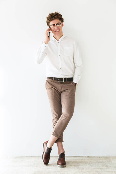 Full length portrait of a successful smiling young attractive curly haired businessman standing infront of white background, talking on mobile phone - 写真・画像