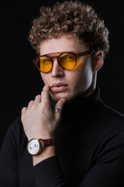 Close up of a handsome confident young curly haired businessman wearing turtleneck standing infront of isolated black background, posing in glasses - Zdjęcie, obraz