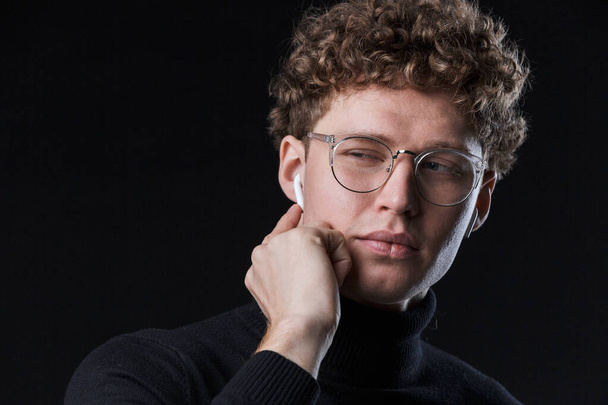 Close up of a handsome confident young curly haired businessman wearing turtleneck standing infront of isolated black background, listening to music with wireless earphones - Фото, изображение