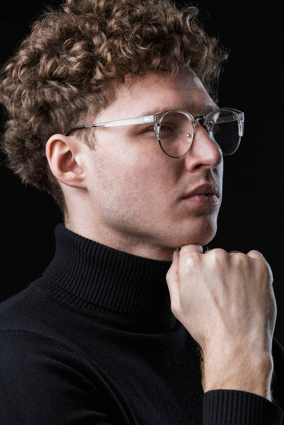 Close up of a handsome confident young curly haired businessman wearing turtleneck standing infront of isolated black background, posing in glasses - Foto, Imagem