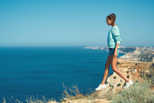 Full length portrait of young slim woman taking step above the cliff, standing in front beautiful sea view. - Foto, imagen