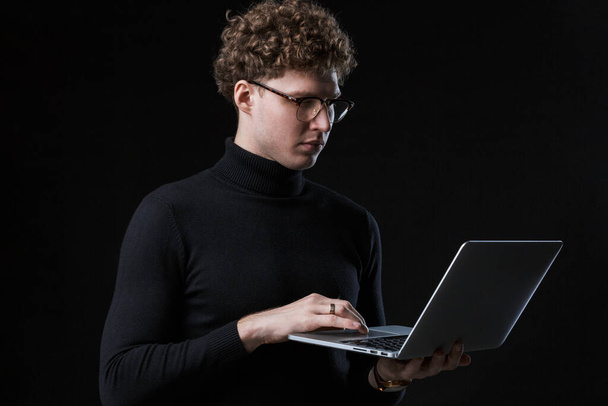 Handsome confident young curly haired businessman wearing turtleneck standing infront of isolated background, working on laptop computer - 写真・画像