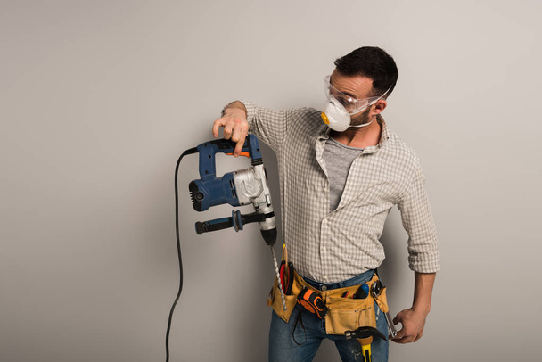 удивленный ручной рабочий в защитной маске держит электрическую дрель на сером
 - Фото, изображение