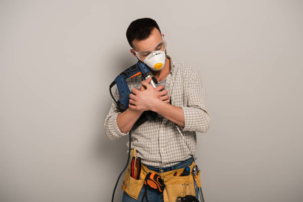 trabalhador manual com olhos fechados em máscara de segurança abraçando broca elétrica em cinza
 - Foto, Imagem