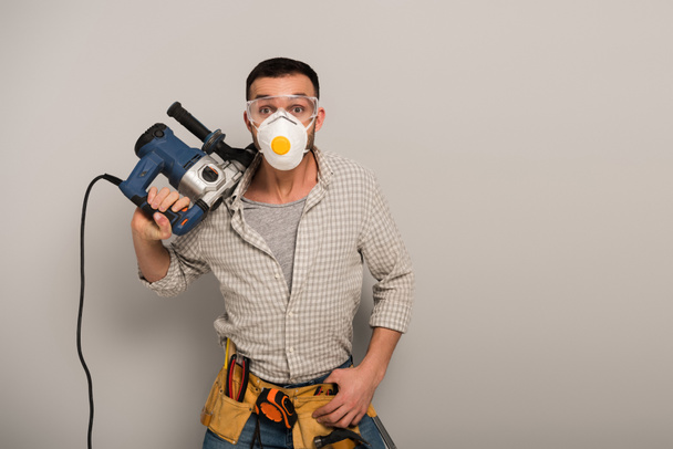 emotional manual worker in safety mask with tool belt holding electric drill on grey - Foto, afbeelding