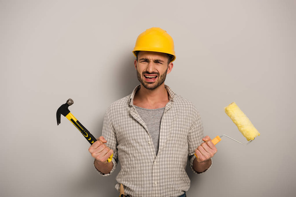 στρεσαρισμένος χειρωνακτικός εργάτης σε σκληρό καπέλο κρατώντας ρολό χρώματος και σφυρί σε γκρι  - Φωτογραφία, εικόνα