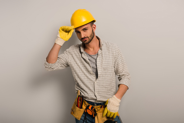 впевнений ремонтник у шоломі та рукавичках з поясом інструментів на сірому
   - Фото, зображення