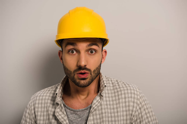 handsome shocked manual worker in yellow hardhat on grey  - Fotografie, Obrázek
