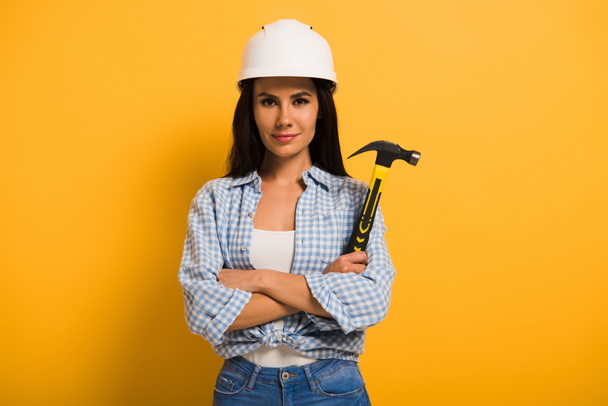 confident female manual worker in helmet holding hammer with crossed arms on yellow  - Photo, image