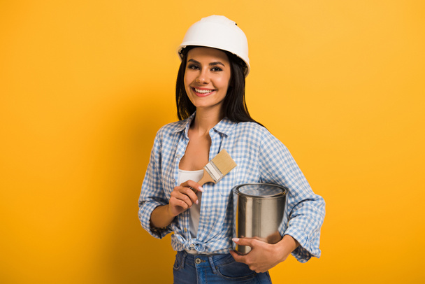 cheerful workwoman in helmet holding paint can and brush on yellow  - Fotografie, Obrázek