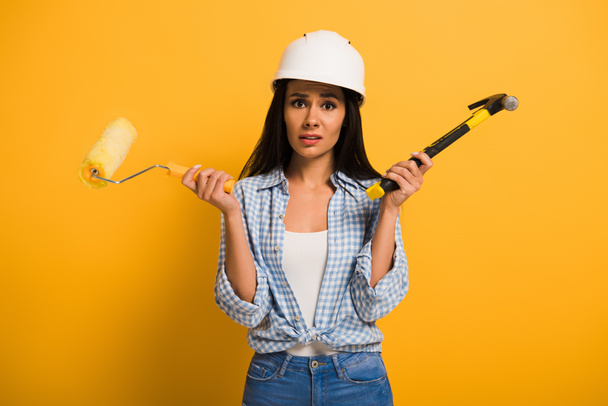 trabajadora frustrada en casco sosteniendo martillo y rodillo de pintura en amarillo
  - Foto, Imagen