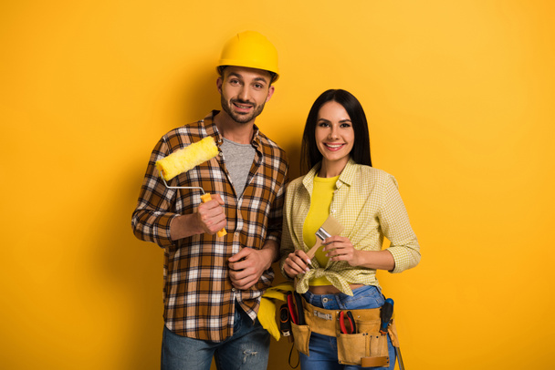professional smiling manual workers holding paint roller and brush on yellow      - Photo, Image