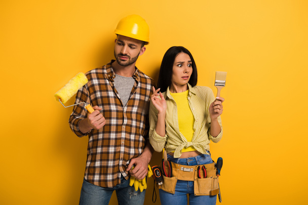 trabajadores manuales confundidos mirando rodillo de pintura y pincel en amarillo
      - Foto, Imagen
