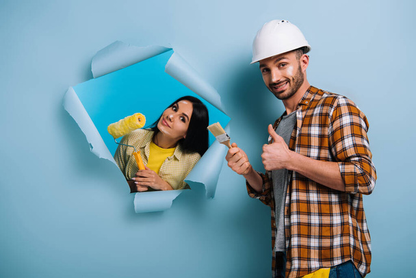 uśmiechnięty pracownik z pędzlem do malowania pokazującym kciuk w górze i kobieta z wałkiem do malowania w podartym papierze, odizolowana na niebiesko - Zdjęcie, obraz